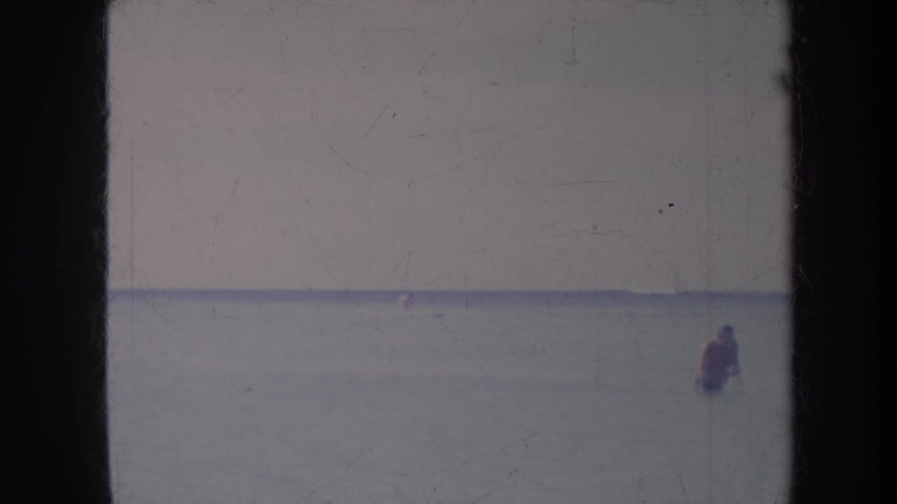 Distant person standing in calm ocean water below a pale sky.