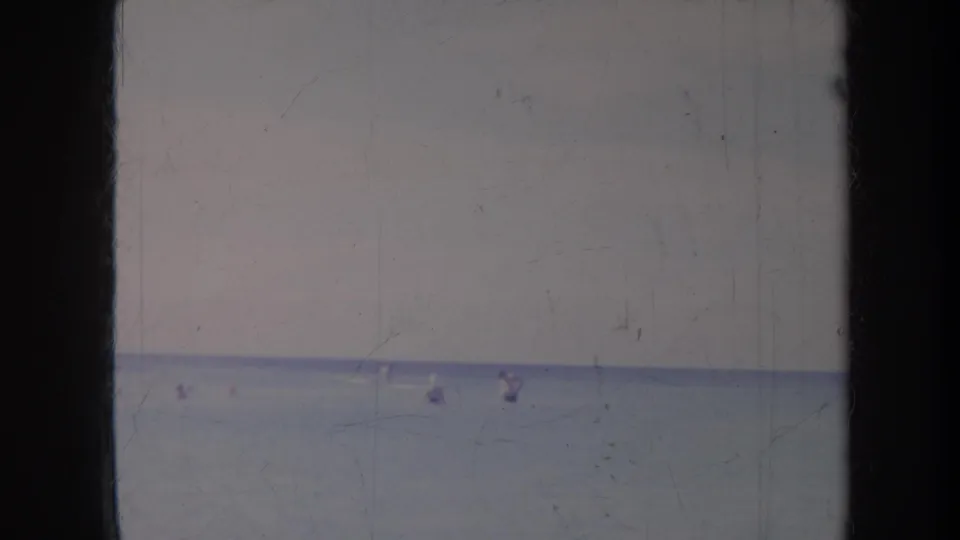 1967 Honolulu ocean scene with several distant figures wading or playing in shallow water beneath a pale sky.