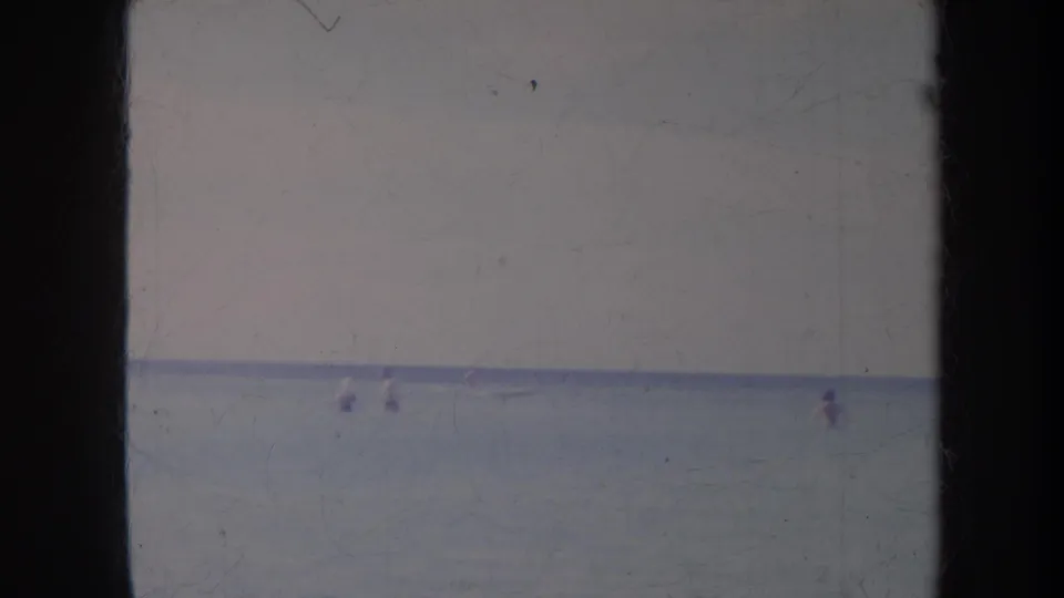 1967 Honolulu home-movie frame showing distant people in calm ocean water with faded blue tones and visible film wear.