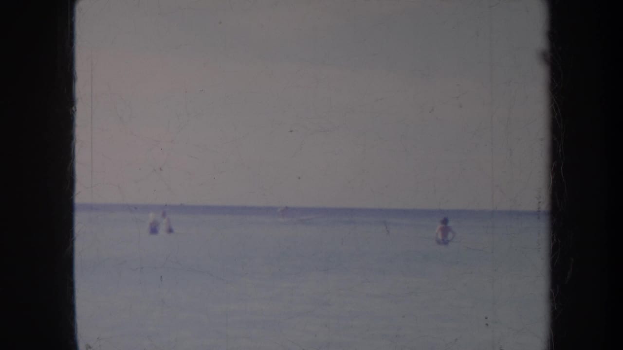 1967 Honolulu home-movie frame with distant figures in calm ocean water under a soft pale sky.