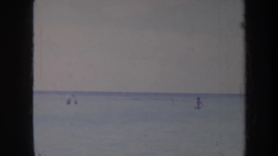 1967 Honolulu home-movie frame with distant figures in calm ocean water under a soft pale sky.