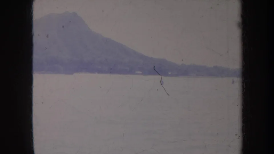 1967 Honolulu home-movie frame showing a distant mountain silhouette across calm coastal water with visible film wear.