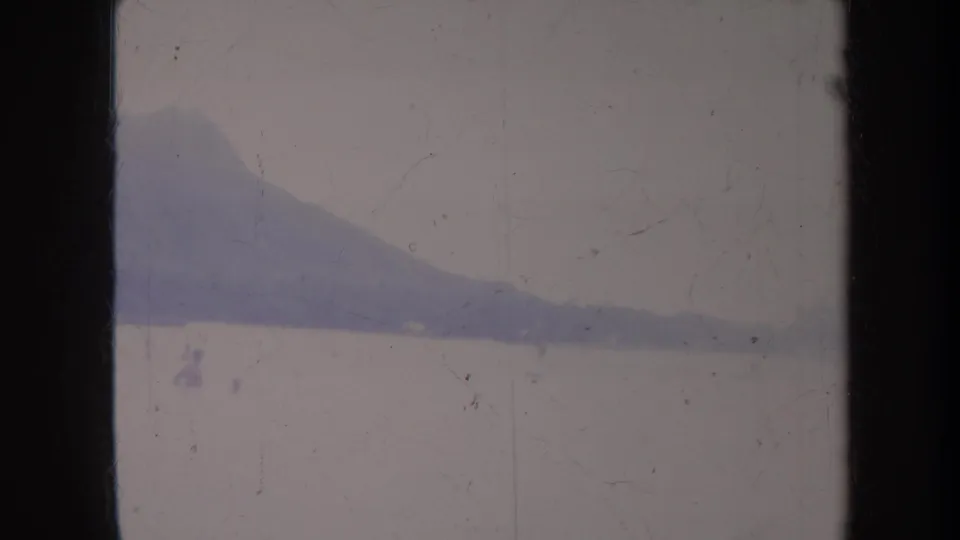 Honolulu 1967 home-movie frame showing a hazy mountain silhouette over calm coastal water with aged film texture.
