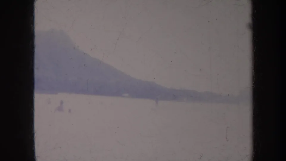 1967 Honolulu home-movie frame showing a hazy mountain ridge beyond calm water with small distant activity on the surface.
