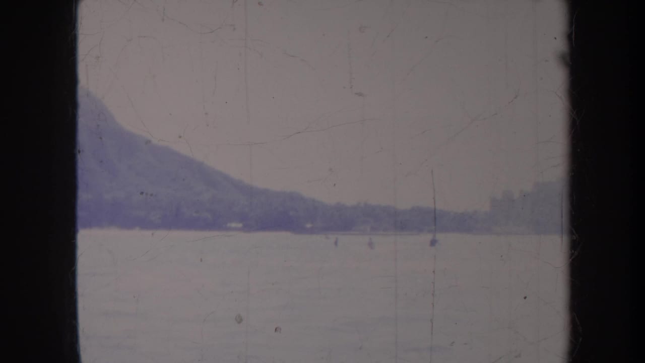 Mountain slope and open water seen through faded, scratched film.