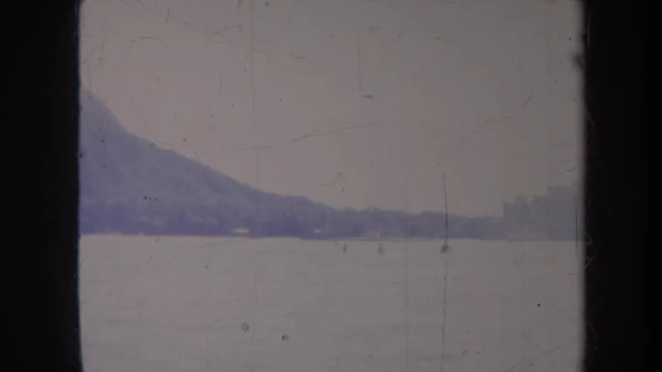 Faded 8mm frame from Honolulu in 1967 with a mountain slope, broad water, and small distant water activity.