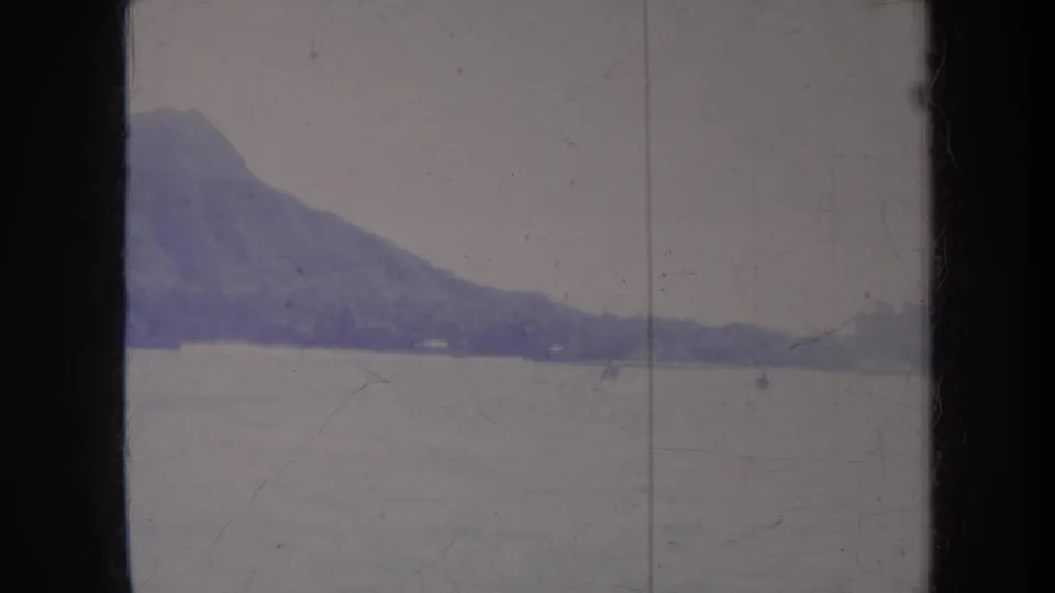 Soft, faded Honolulu 1967 home-movie frame of a mountain ridge meeting calm water with distant recreation barely visible.