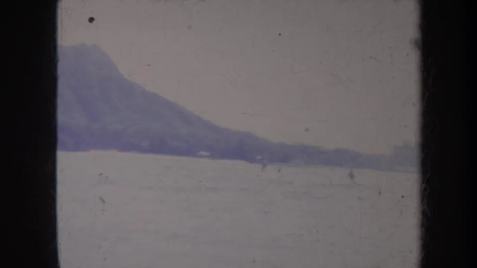 1967 Honolulu 8mm frame showing open water, a sloping mountain ridge, and tiny distant figures or craft near the horizon.