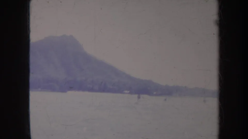 1967 Honolulu 8mm frame looking across washed-out ocean water toward Diamond Head with small distant recreation figures.