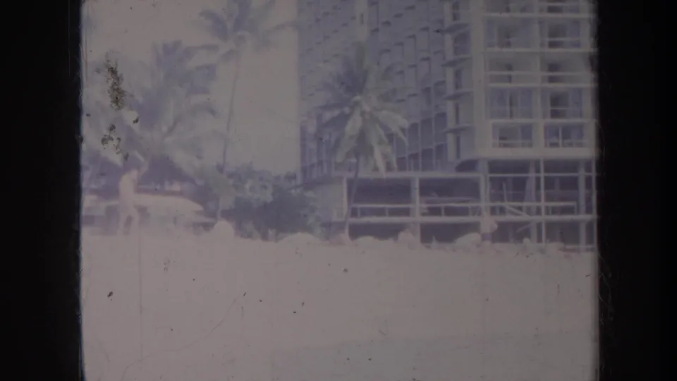 1967 Honolulu home-movie frame showing palms and a multi-story resort building near the beach in faded color.
