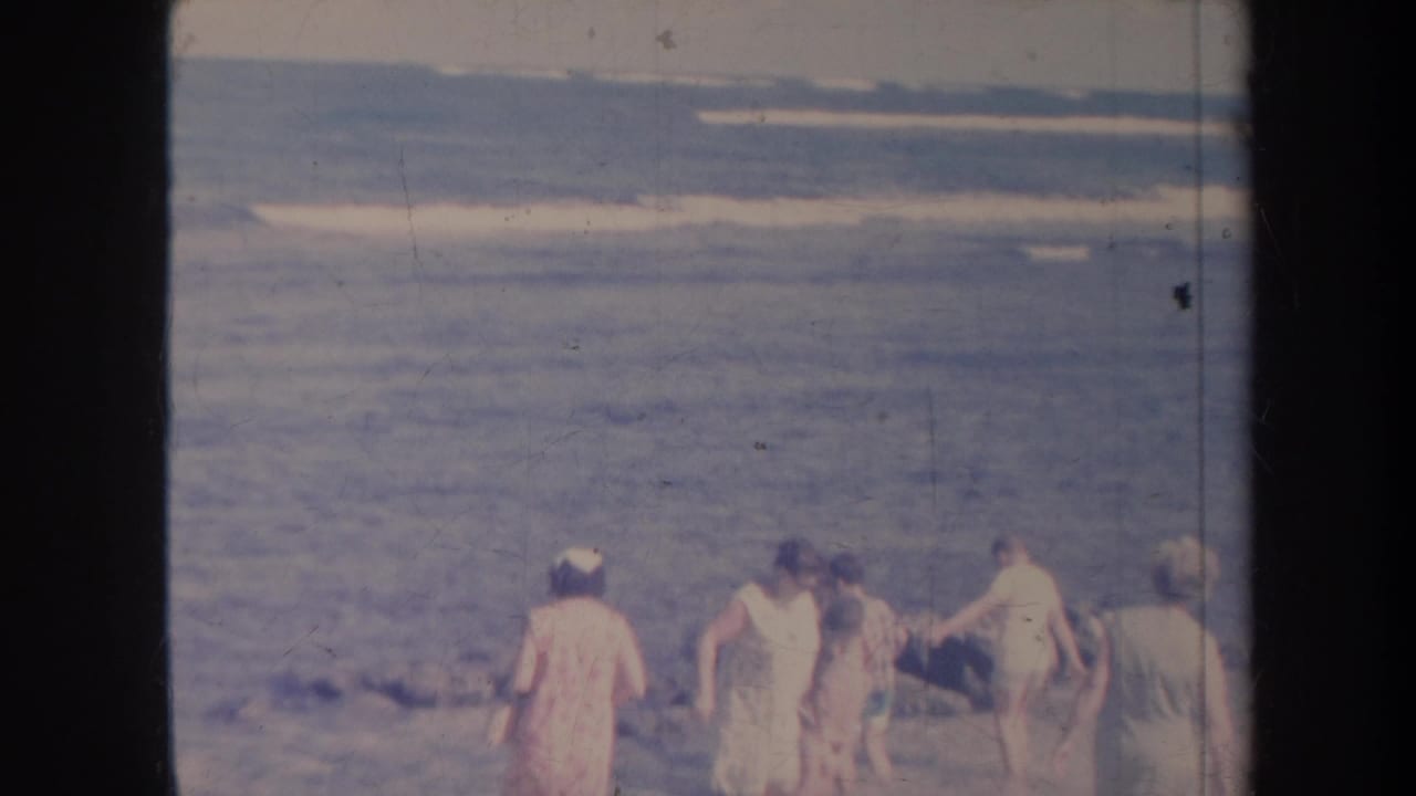 People in beachwear walk near ocean surf on a rocky shoreline.