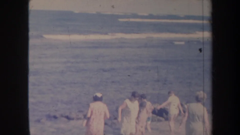 1967 Honolulu home-movie frame showing people in period beachwear near waves on a rocky shore.