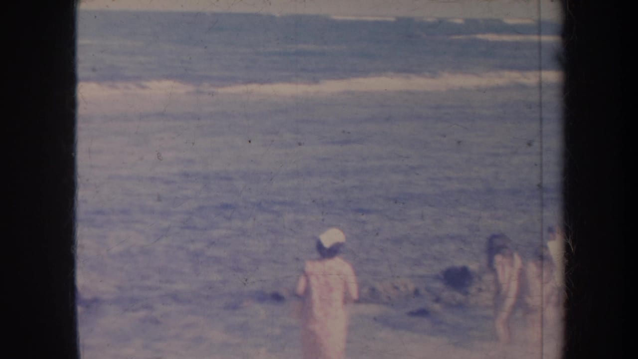 1967 Honolulu shoreline frame with surf, a person in a light cap, and partially visible beachgoers at the edge of the scene.