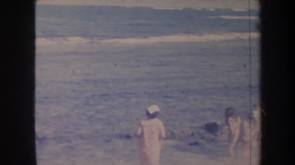 1967 Honolulu shoreline frame with surf, a person in a light cap, and partially visible beachgoers at the edge of the scene.