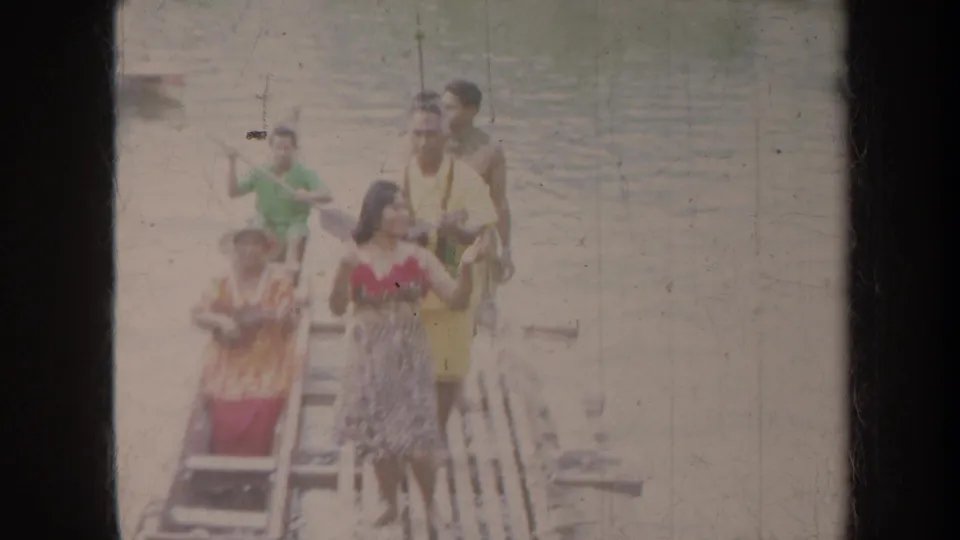 1967 Honolulu home-movie frame showing people on a wooden dock, including a woman in a floral top and others with paddles or leis.