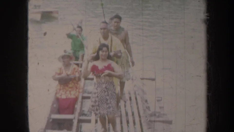 1967 Honolulu frame of dock performers facing the camera, with paddles, leis or garlands, and water behind them.