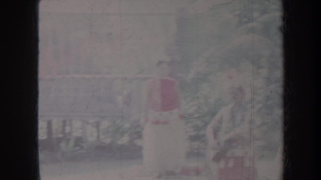 Overexposed hula dancer in red and white costume near a porch railing and palm foliage.