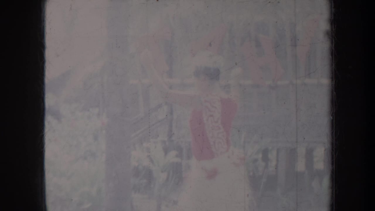 Dancer with red garment raises an arm in a tropical outdoor setting.