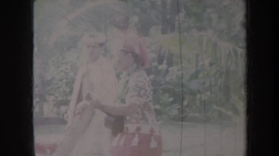 1967 Honolulu frame showing a dancer in pale dress with red floral details near palm fronds and garden foliage.