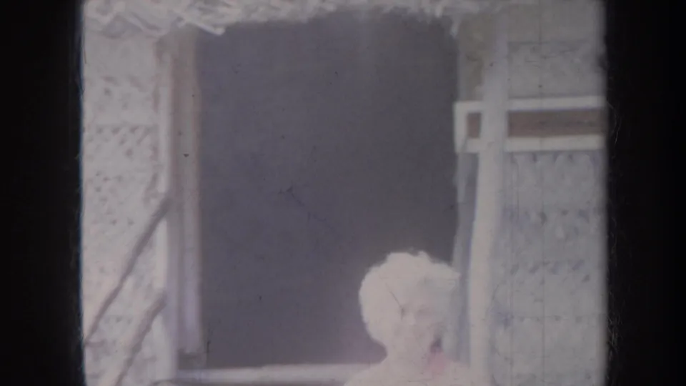 1967 Honolulu home-movie frame showing an adult with short light hair near a thatched roof edge and woven wall panels.