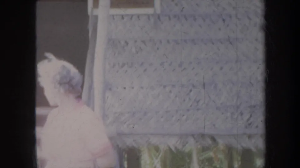 1967 Honolulu frame of a woman in a light dress near lattice wall panels and a shaded thatched setting.