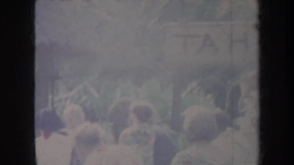 A Honolulu resort crowd is seen from behind in soft 8mm footage, with heads, hats, foliage, and a thatched structure visible.