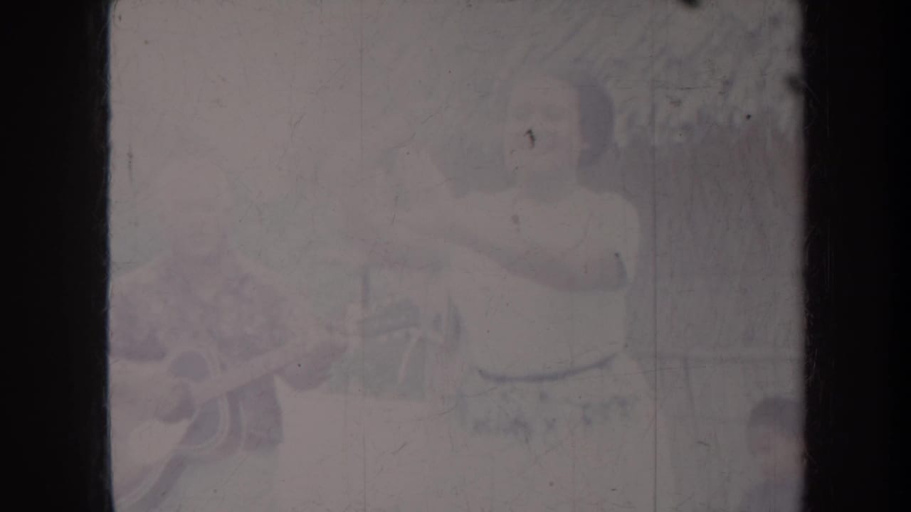 1967 Honolulu frame with a woman gesturing in dance beside a seated acoustic guitarist in a casual outdoor scene.