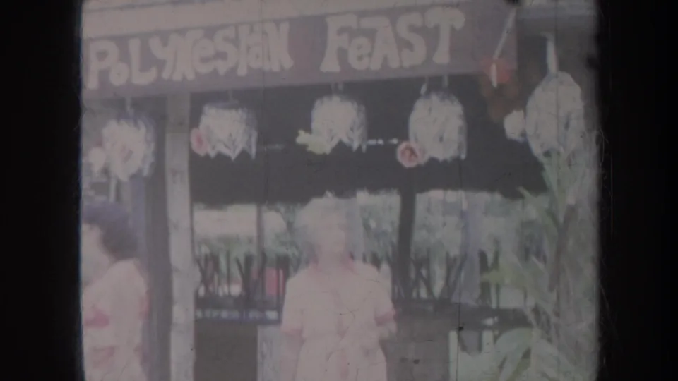 1967 Honolulu travel frame showing visitors near storefront windows under a sign reading Polynesian Feast.
