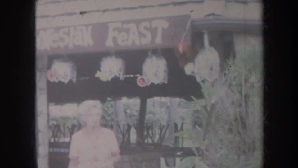 1967 Honolulu home-movie frame showing a woman near a Polynesian Feast storefront with hanging floral decorations.