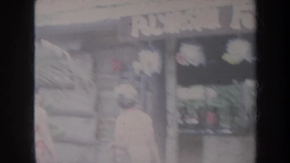 Soft 1967 Honolulu street frame with a woman walking past tropical plants and a storefront sign.