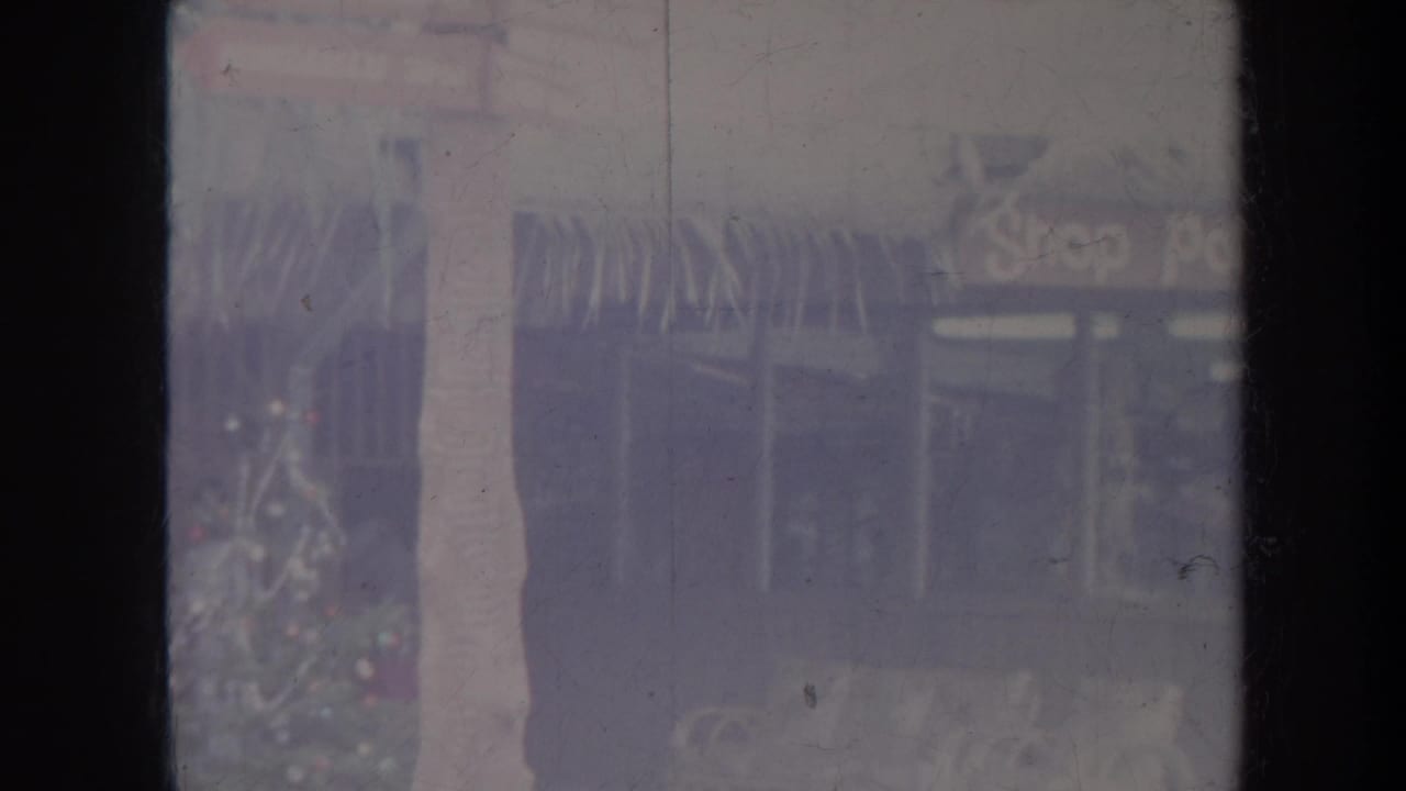 1967 Honolulu home-movie frame with storefront windows, a signpost, and tropical planting in soft faded color.