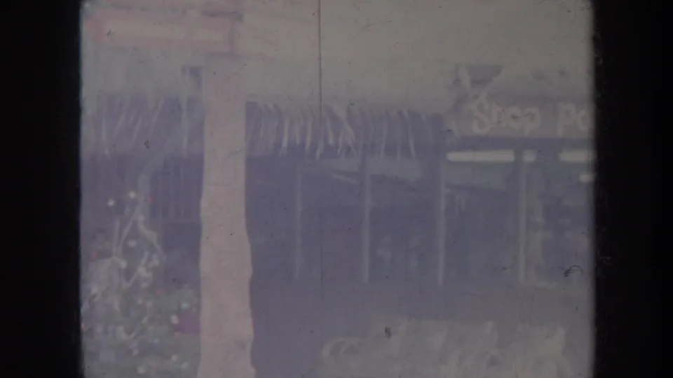 1967 Honolulu home-movie frame with storefront windows, a signpost, and tropical planting in soft faded color.