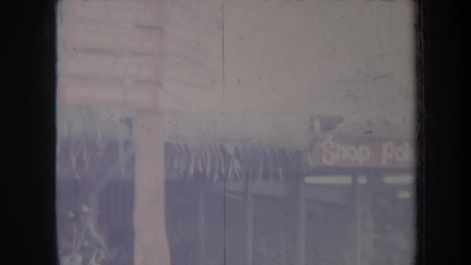 1967 8mm travel frame showing a hazy tropical storefront with palms, partial shop signage, and heavy surface scratches.