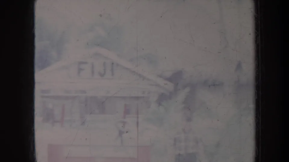 1967 Honolulu home-movie frame showing a FIJI-painted hut facade behind palm foliage, with faded color and visible film wear.