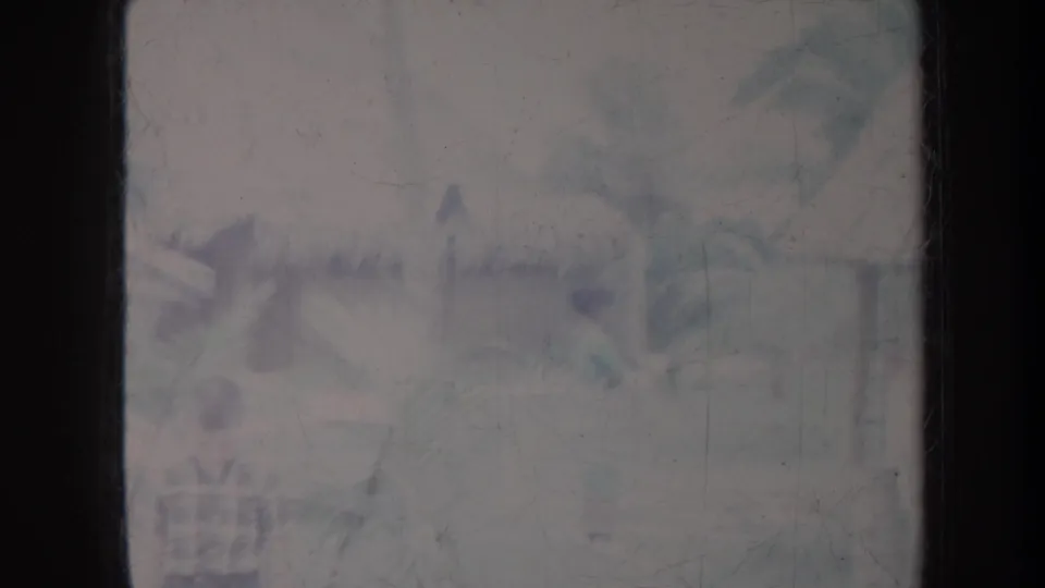 1967 Honolulu home-movie frame of a tropical village area with palms, a distant person, and indistinct hut or roofline forms.