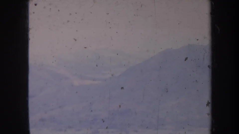1967 Honolulu home-movie frame with hazy mountain slopes and pale sky, marked by grain, dust specks, scratches, and film gate edges.