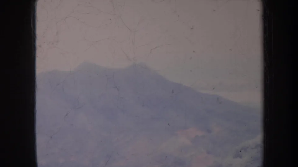 1967 Honolulu home-movie frame of a distant hazy mountain range, rendered in muted color with visible dust, scratches, grain, and gate edge.