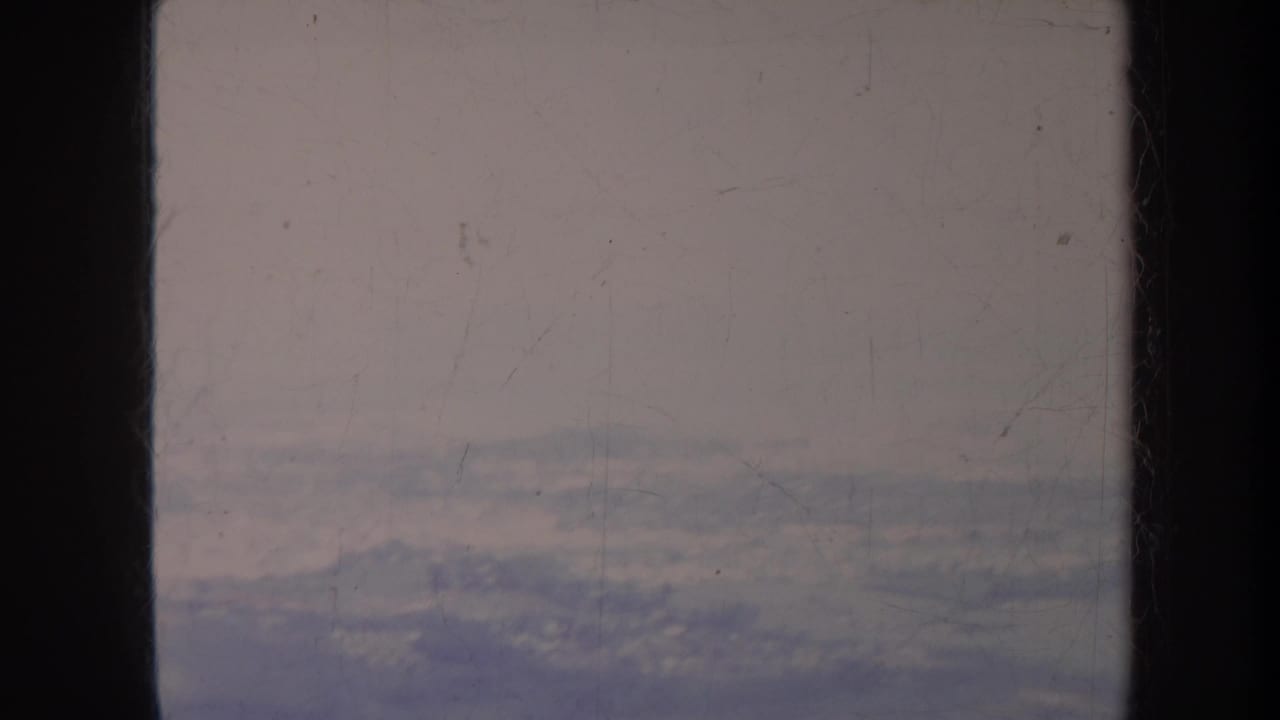 Aerial view through dark aircraft-window framing with cloud cover and faint land below.