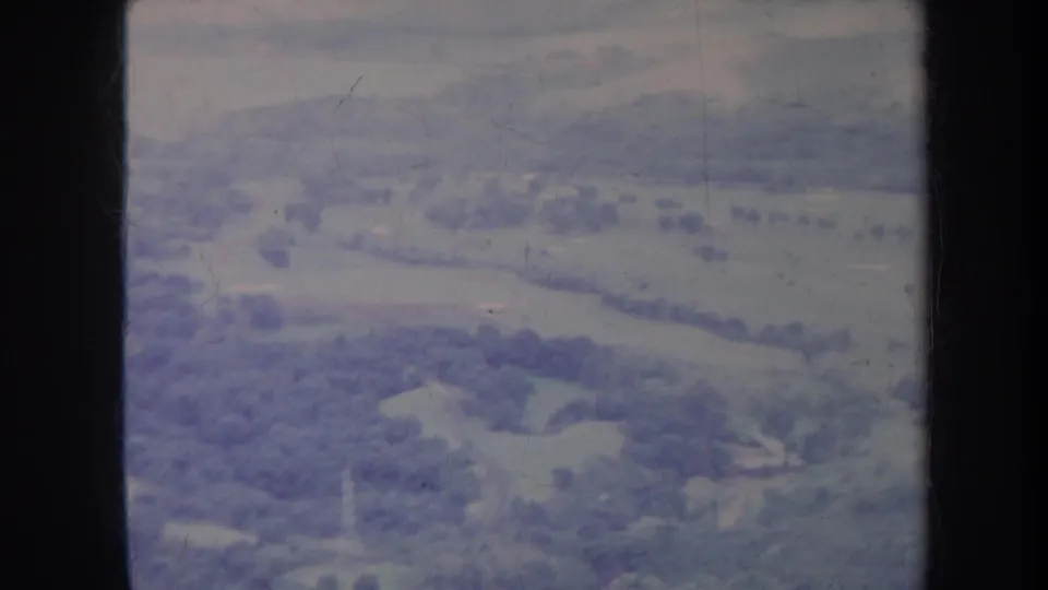 1967 Honolulu-area aerial home-movie frame showing open rolling terrain, sparse foliage, and soft blue-purple 8mm color.
