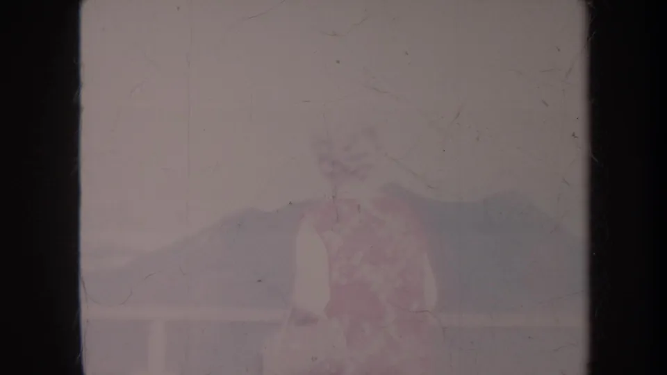1967 Honolulu home-movie frame showing a woman in a floral dress near a balcony or overlook with distant hills.