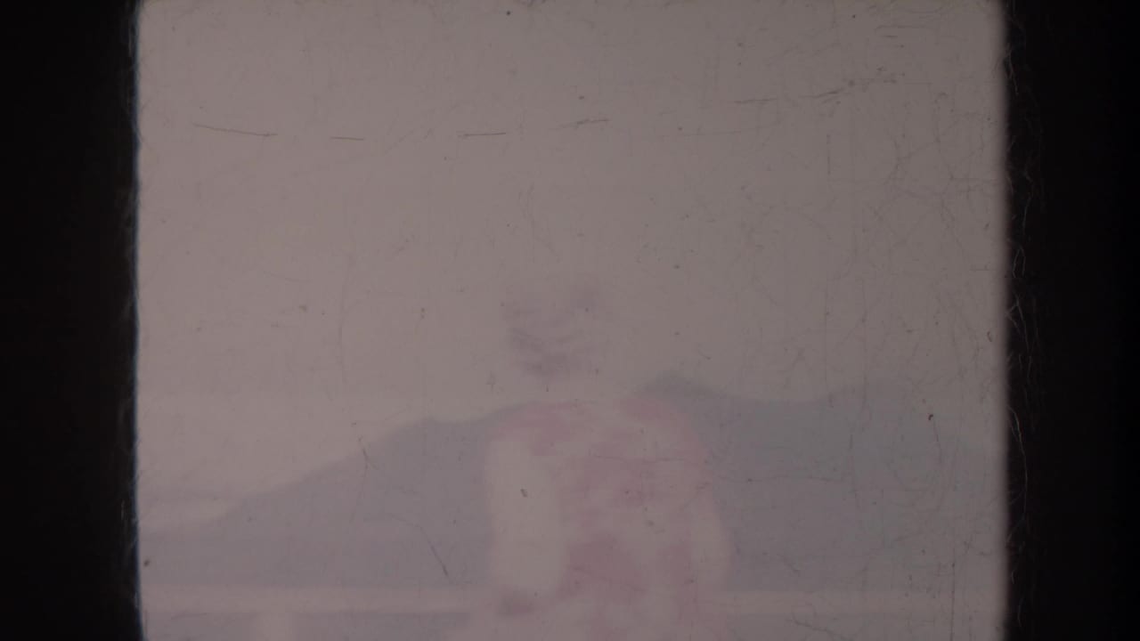 Faded coastal view with dark film borders, pale sky, distant shoreline, and faint silhouettes.