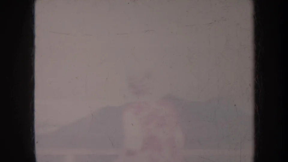 1967 Honolulu 8mm frame with a hazy seaside backdrop, dark film borders, faded color, and faint distant figures near the shore.