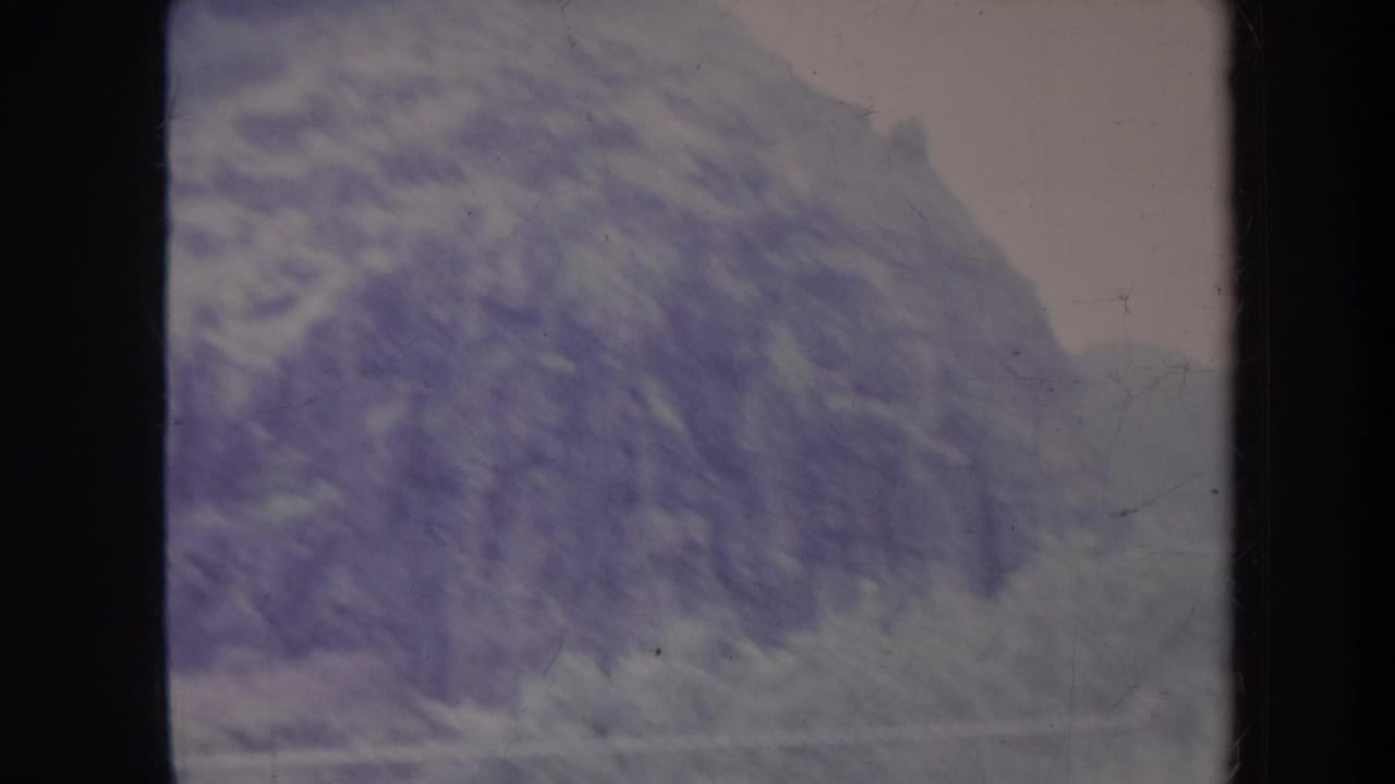 1967 8mm travel footage frame showing a fog-softened mountain slope, dark gate edges, and faded color tones.