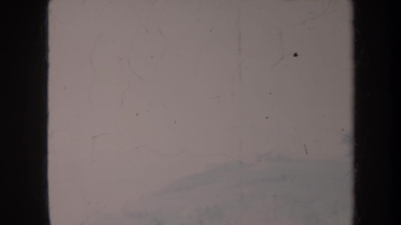 Faded film frame with dark vertical edges and a hazy hillside barely visible.