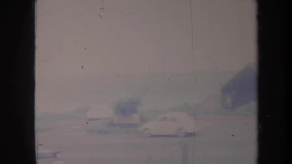 Blurred 1967 Honolulu roadside frame with cars, a low structure, pale sky, and visible 8mm wear.