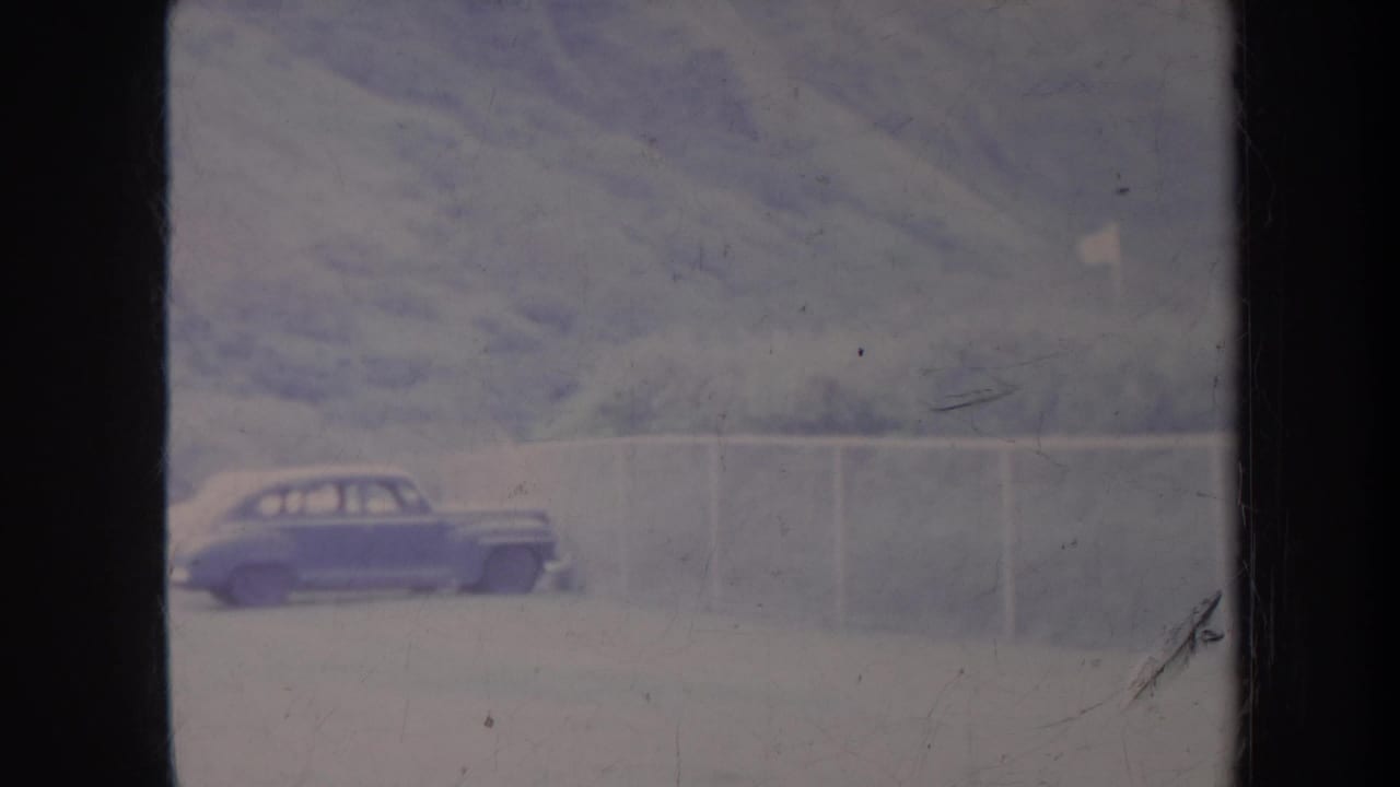 Blue car parked near a pale fence with green hills behind it.
