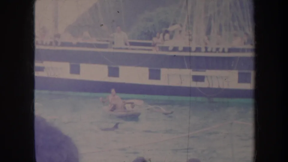 Faded 1967 Honolulu home-movie frame showing people around a small raft with a larger vessel across the background.