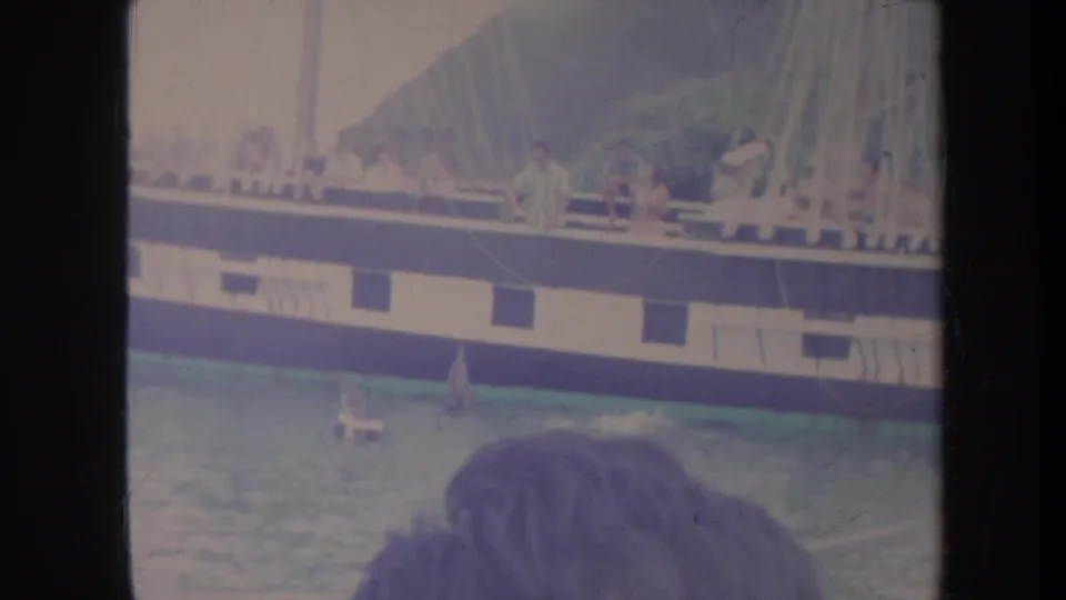 1967 Honolulu 8mm frame with dolphins near a sailboat, passengers on deck, rigging above, and tropical hillside beyond.