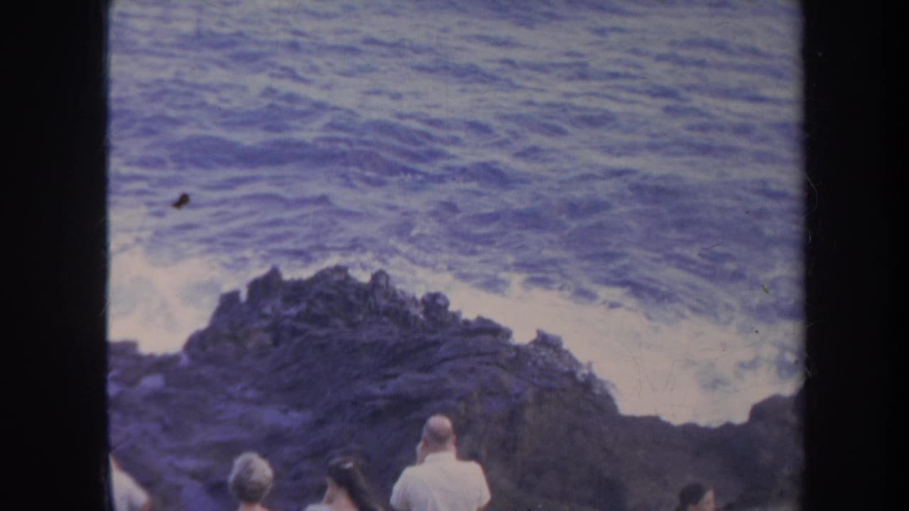 Tourists stand near a rocky shore as rough ocean water breaks behind dark rocks.