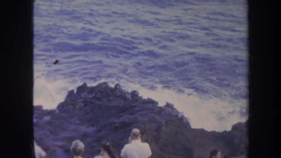 1967 Honolulu home-movie frame showing visitors from behind watching waves hit dark shoreline rocks.